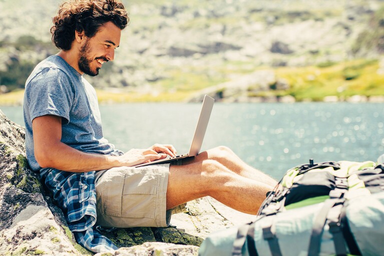 Mann mit einem Laptop auf den Knien sitzt an einem Bergsee