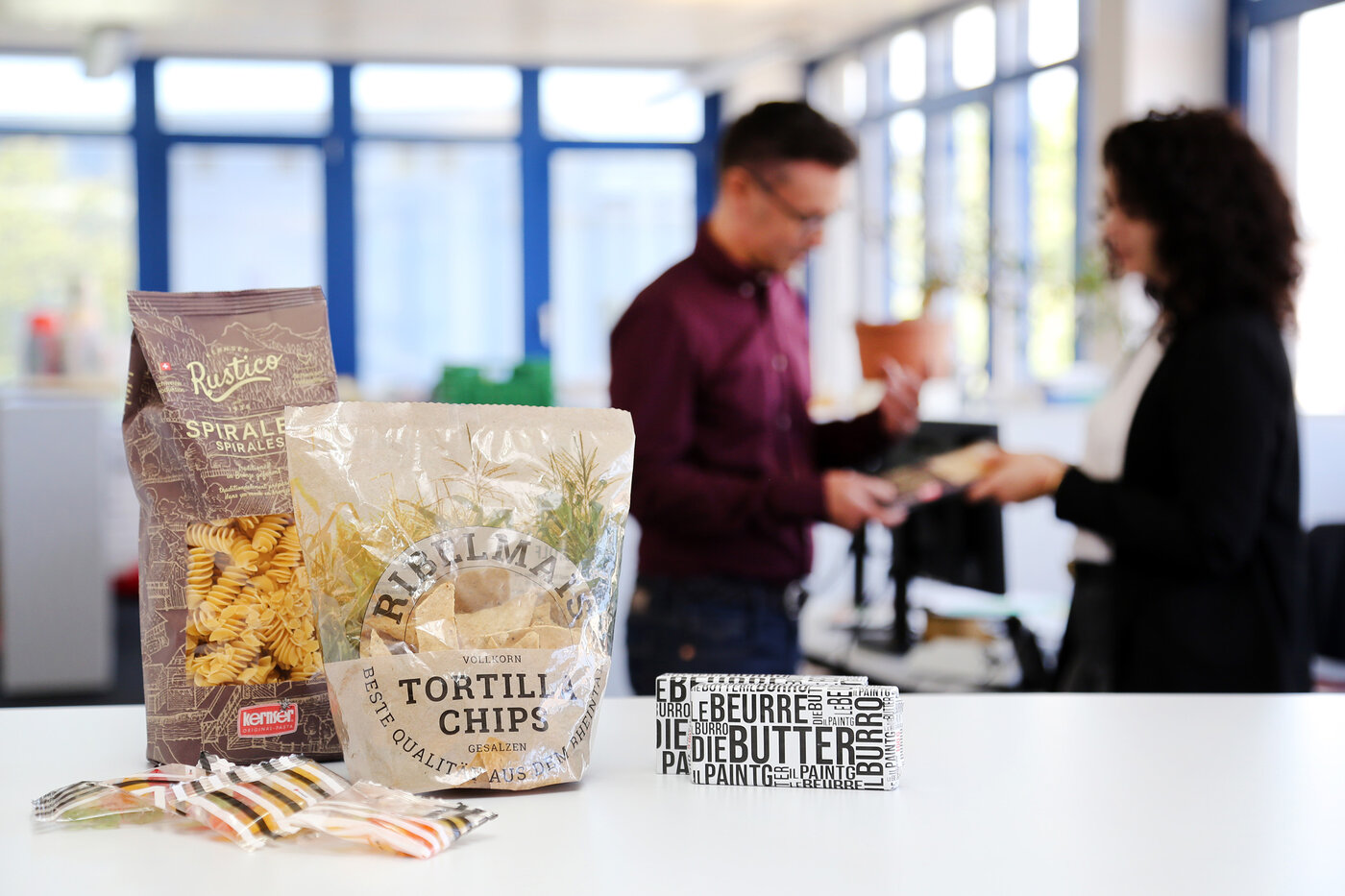 Im Vordergrund Prototypen von Beuteln, Sachets und Butterwickelfolien. Im Hintergrund zwei Personen im Gespräch. 