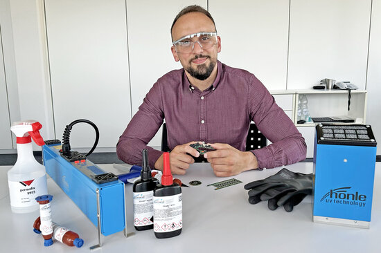 Ein Permapack-Mitarbeiter sitzt an einem Tisch, auf dem UV-Klebstoffe, Schutzhandschuhe und ein UV-Gerät platziert sind.