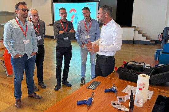 Eine Gruppe Kunden steht mit einem Permapack-Mitarbeiter vor einem Tisch mit Klebeutensilien.