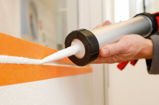 Ein Arbeiter spritzt Dichtmasse aus einer Handpresspistole in eine mit orangen Abdeckband abgeklebte Fuge. 