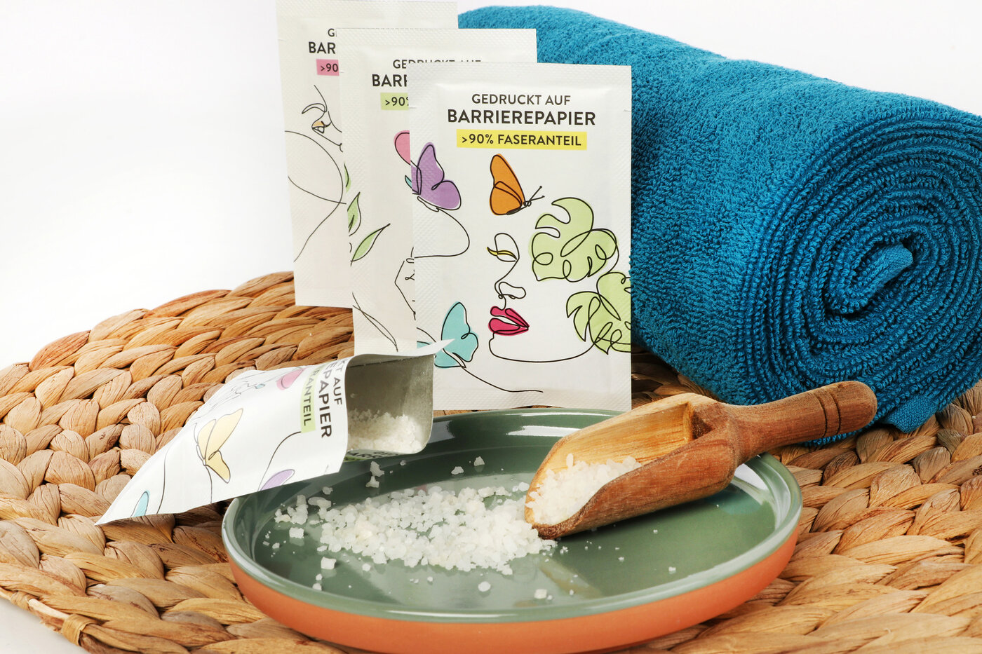 Drei Barrierepapier-Verpackungen mit Badesalz lehnen an einer Handtuchrolle.
