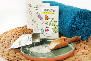 Drei Barrierepapier-Verpackungen mit Badesalz lehnen an einer Handtuchrolle.