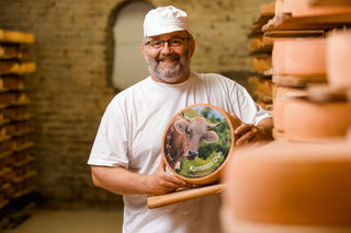 Ein Mann zeigt in einem Käsereifekeller einen runden Käse mit einer bedruckten Käselaibetikette.