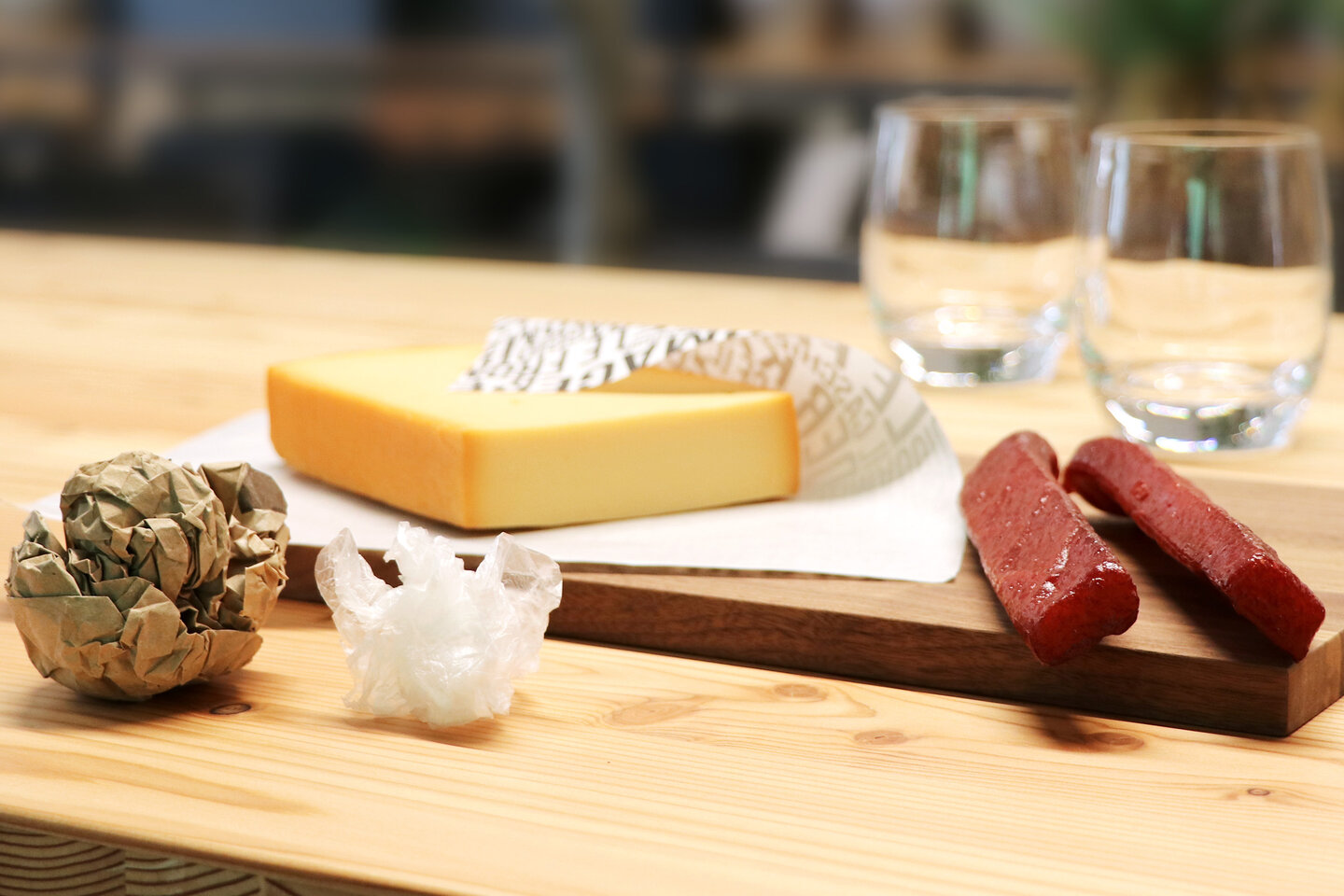 Ein Stück Käse und ein paar Landjäger liegen auf einem Brotzeitbrett, daneben zusammengeknülltes Papier und zusammengeknüllte Folie. 