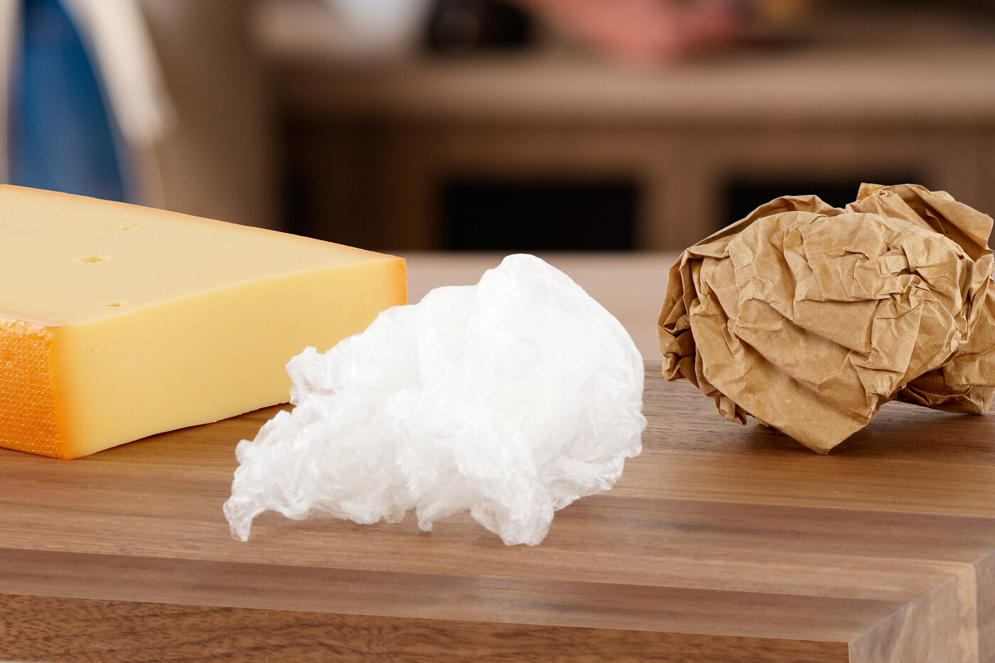 Ein Stück Käse auf einem Brotzeitbrett, daneben zusammengeknülltes Papier und zusammengeknüllte Folie. (