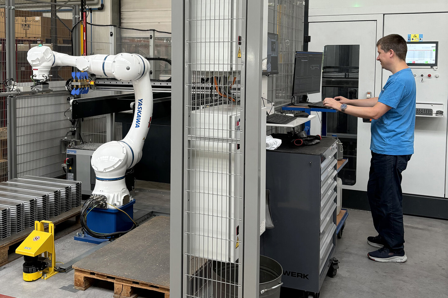 Rechts ein Mitarbeiter am PC; links stapelt ein Roboter zurechtge­schnittene Profile auf einer Palette.