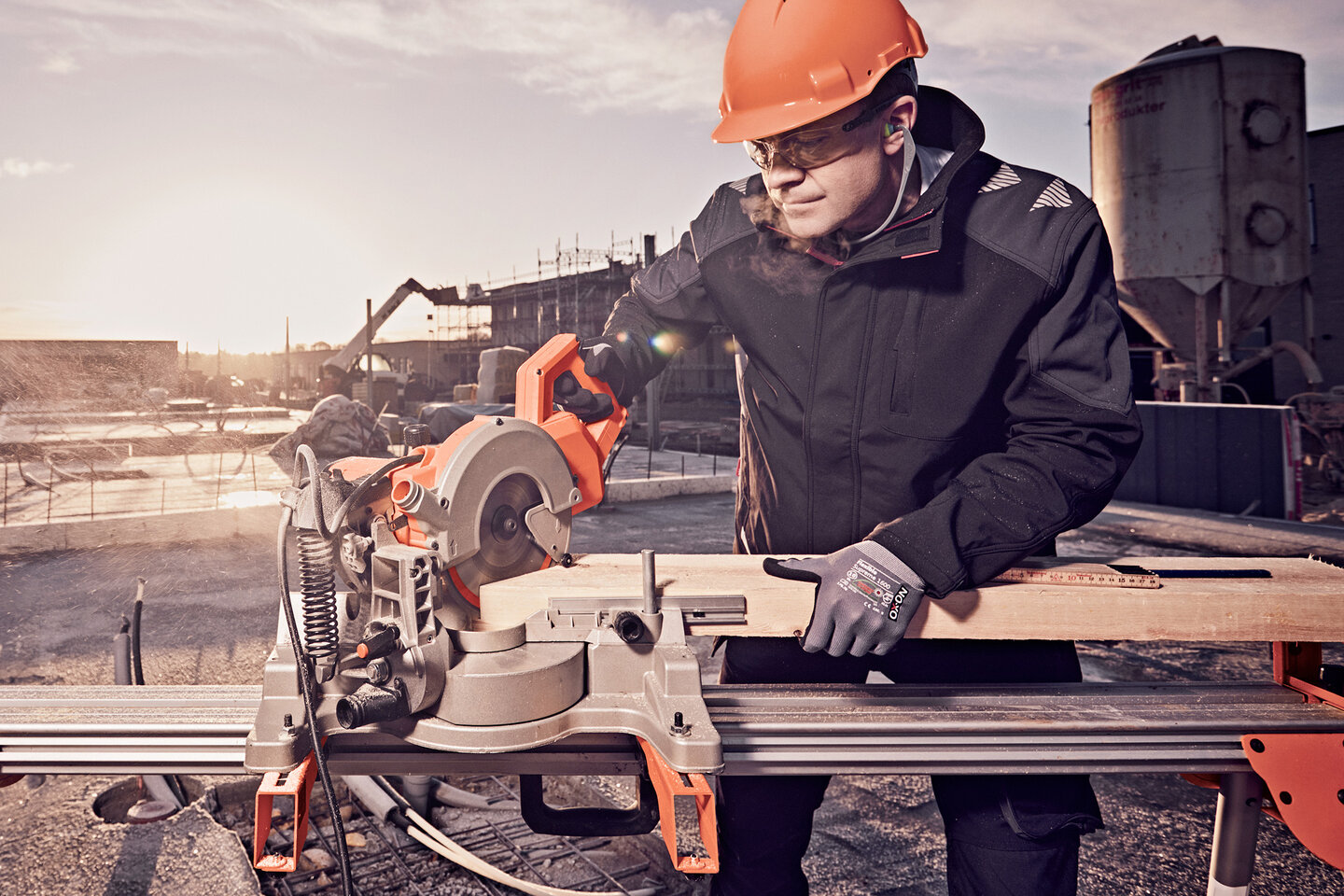 Ein Handwerker auf der Baustelle mit Schutzhelm, Schutzbrille und Schutzhandschuhen bedient eine Kappsäge und schneidet ein Holzbrett zu. 