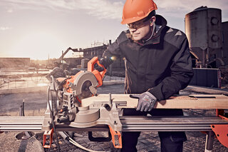 Ein Handwerker auf der Baustelle mit Schutzhelm, Schutzbrille und Schutzhandschuhen bedient eine Kappsäge und schneidet ein Holzbrett zu. 