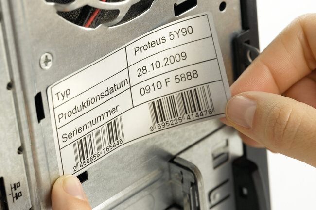 Eine schwarz bedruckte Geräteetikette wird auf ein Gerät aufgeklebt. 