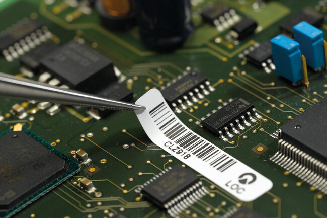 Eine hitzebeständige Etikette wird mit einer Pinzeltte auf eine Leiterplatte geklebt.