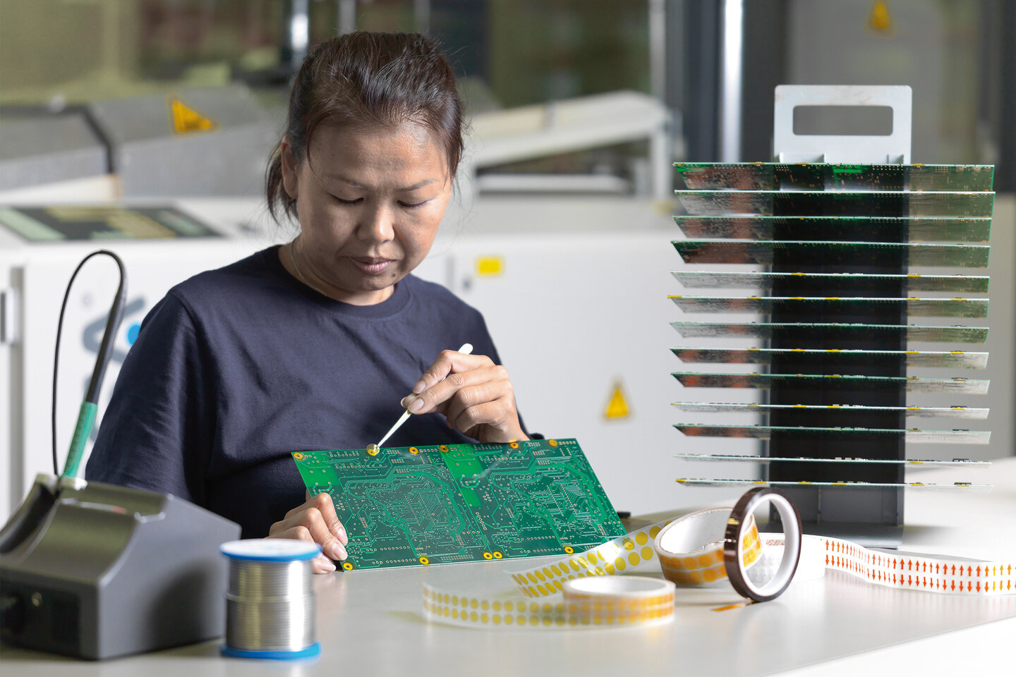 Eine Mitarbeiterin mit einer Pinzette in der Hand klebt Goldkontakte auf einer Leiterplatte mit runden Formstanzteilen ab.