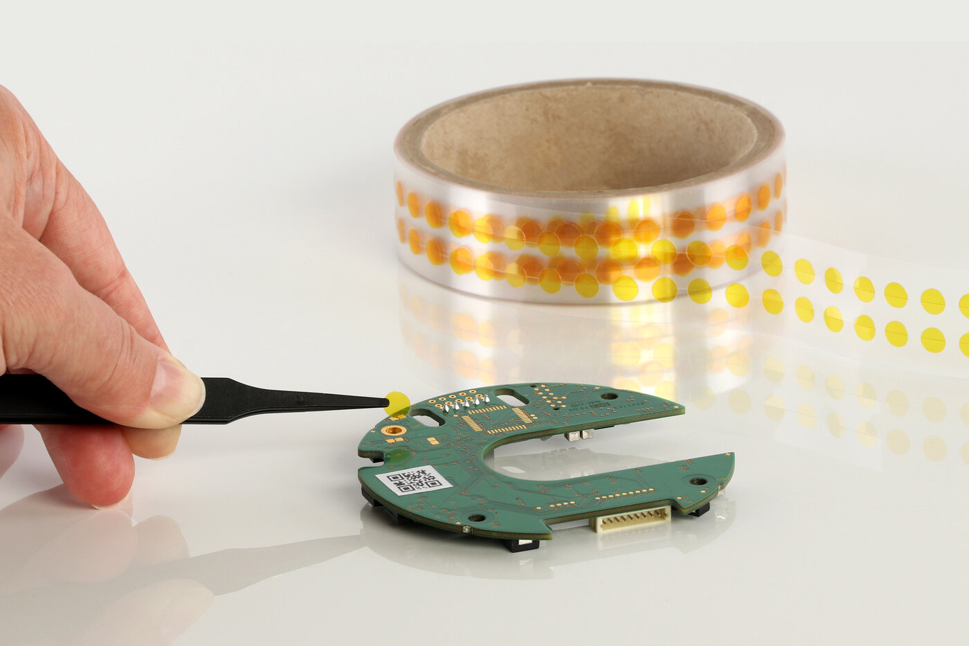 Hand mit Pinzette deckt mit einem kleinen, runden Formstanzteil einen Kontakt auf einer Leiterplatte ab.