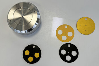 Verschiedene Ebenen eines Ziffernblatts und ein hauchdünnes Klebeband-Formstanzteil liegen nebeneinander. 