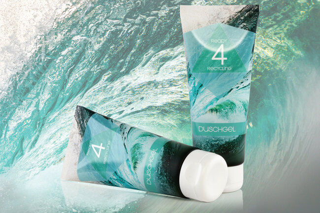Zwei grünblau bedruckte Tuben mit Duschgel aus dem Material Blueloop und im Hintergrund eine sich überschlagende Wasserwelle. 