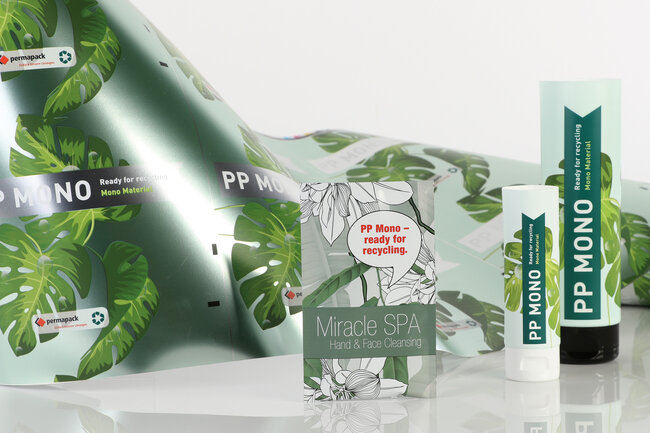 Zwei in Grüntönen bedruckte Tuben und ein Sachet aus dem Material Green Motion. Im Hintergrund eine Rolle mit bedrucktem Mono-Material