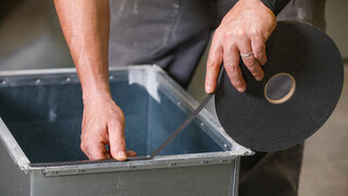 Ein Mann klebt ein schwarzes Dichtungsband auf die Kanten von einem Lüftungsschacht.