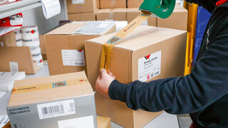 Mann verschliesst einen Karton mit Klebeband, im Vordergrund ein Karton mit einer Dokumententasche.