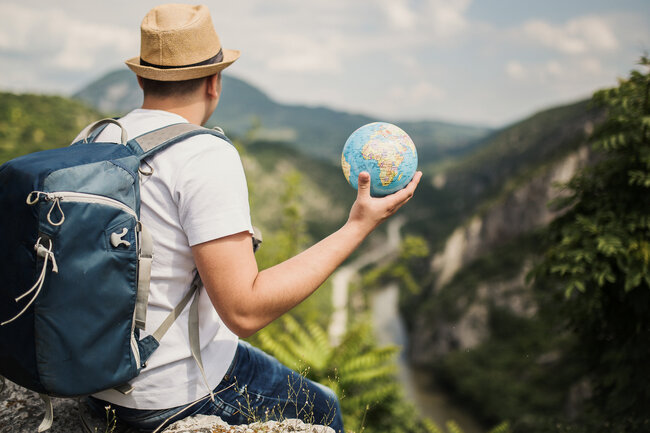 Reisender mit Rucksack hält einen Globus und blickt über eine Berglandschaft
