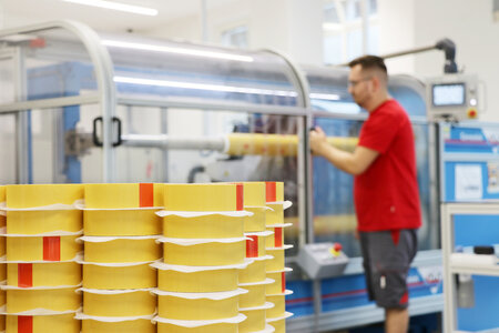 Im Vordergrund ein Stapel mit gelben, geschnittenen Klebebändern. Im Hintergrund ein Mann an einer Maschine. 