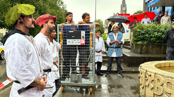 Zwei Männer in einem Gitterwagen und umringt von Mitarbeitenden erwarten die Druckertaufe.
