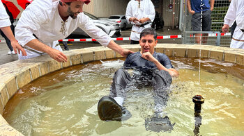 Ein Mann sitzt im mit Wasser gefüllten Brunnen, dahinter vier Männer