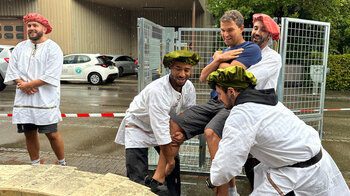 Ein Mann wird von drei Mitarbeitenden hochgehoben, um in den mit Wasser gefüllten Brunnen zu tauchen.
