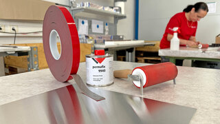 Montageklebeband, Primer und Andrückrolle auf einem Arbeitstisch, im Hintergrund arbeitet eine Frau an einem Werkstück.