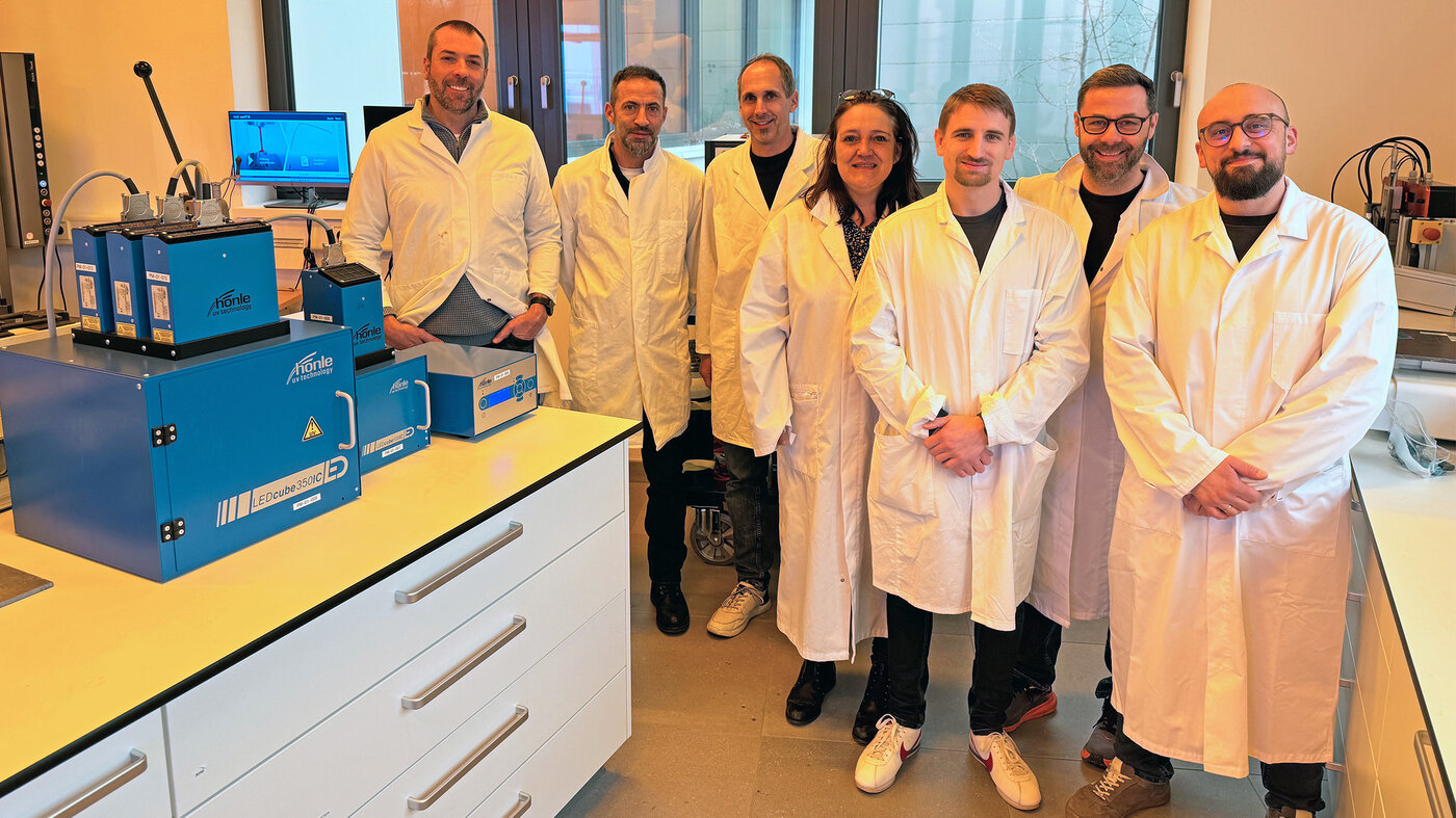 Gruppe von Mitarbeitenden in Laborkitteln. Im Vordergrund stehen UV-Härtungsgeräte und Laborequipment für Klebstofftests.