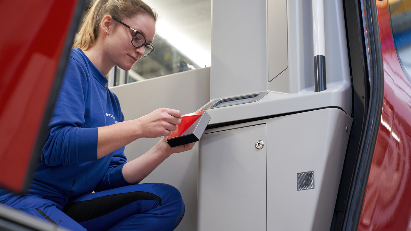 Monteurin kniet in einem Zugabteil und bringt ein Bauteil mit einem Montageklebeband an.