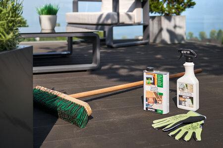 Holzterrasse von der Sonne beschienen, ein Besen zwei Reinigungsmittel und ein Paar Gartenhandschuhe auf dem Boden arrangiert